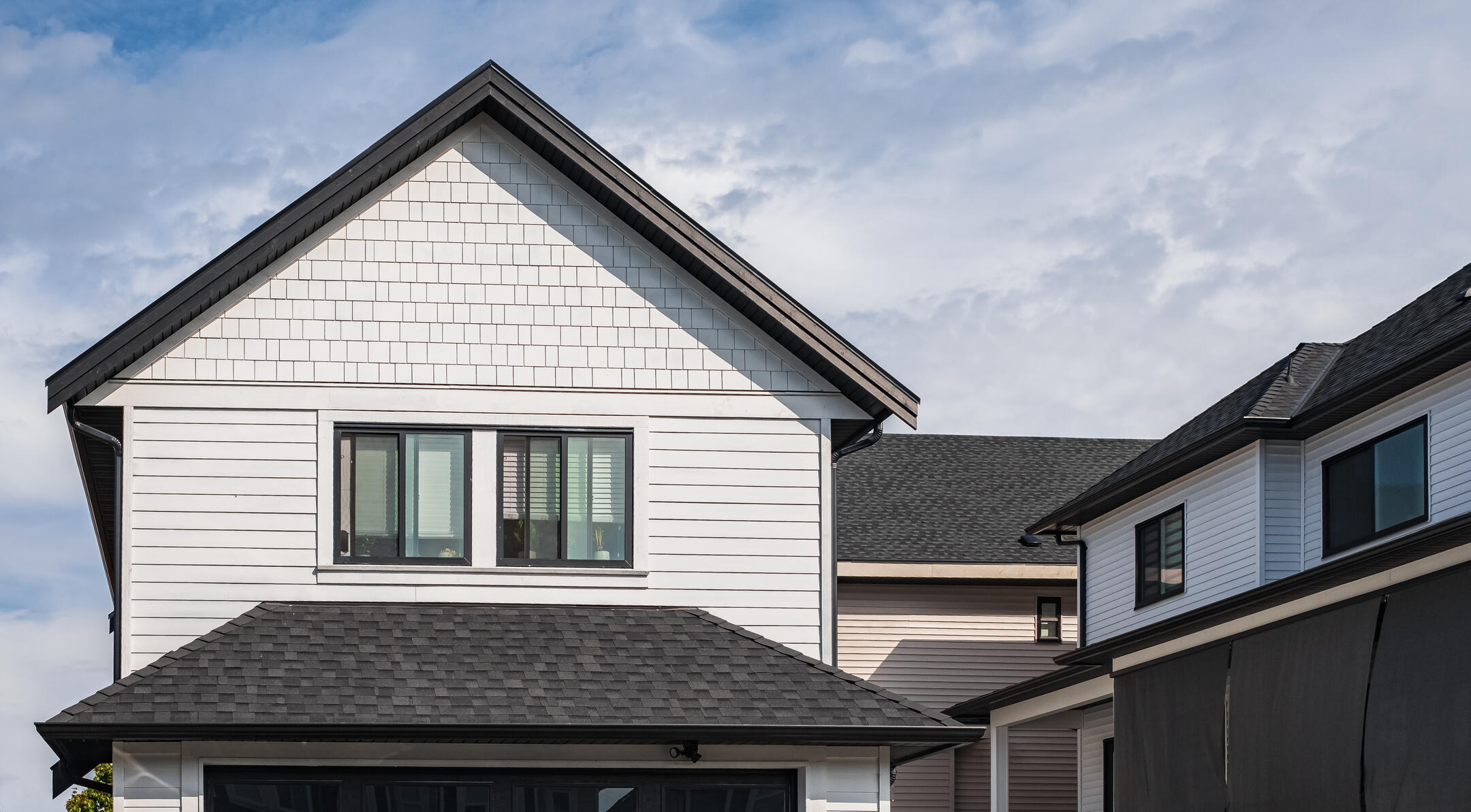 A modern, two-story house with a steep, dark gray roof and white siding, featuring large windows. Another house is partially visible next to it. The sky above is cloudy with patches of blue.