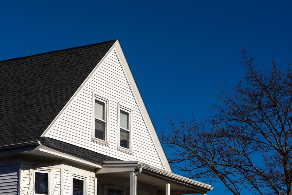 A white house with a steeply pitched roof and two windows on the upper floor. A bare tree without leaves stands to the right, all under a clear blue sky.