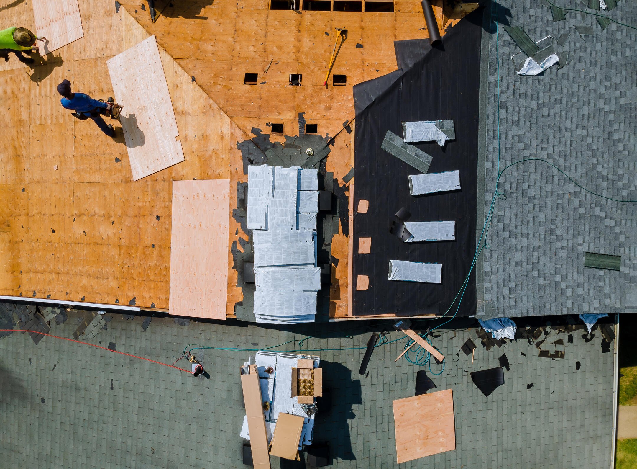 Aerial view of a roof under renovation. Two workers are placing plywood and tar paper, with tools and materials scattered around. Shingles are partially installed on the right side. A ladder leans against the roof.