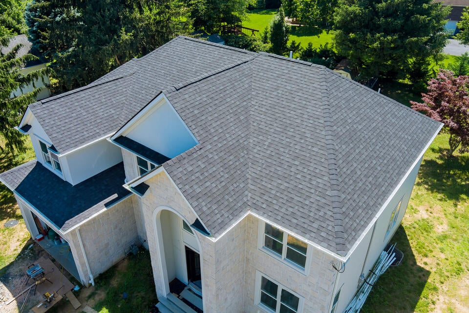 Aerial view of a two-story house with a dark gray shingle roof and light stone exterior. The house features an arched entrance and multiple windows. Surrounding the house is a green lawn with trees and shrubs.