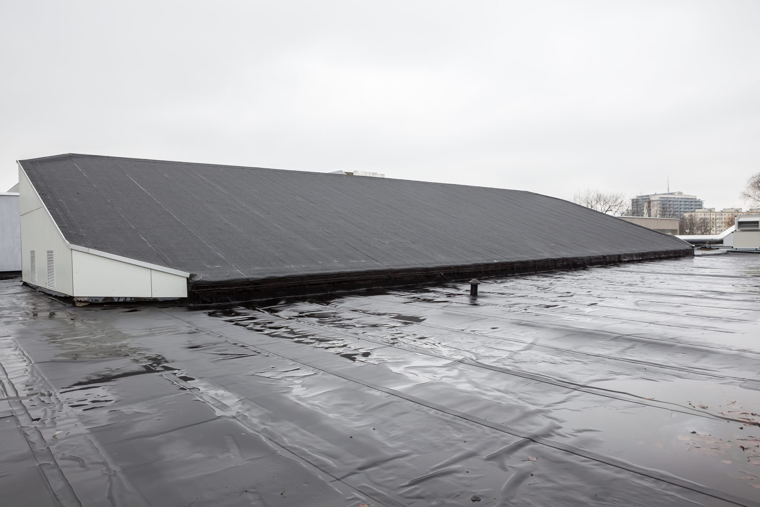 Flat rooftop covered with black waterproof material. A large, pitched structure is centered on the roof. The sky is overcast, and a few distant buildings are visible on the horizon.