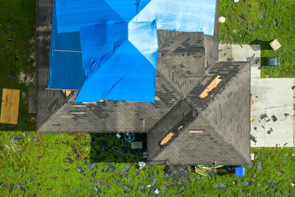 Aerial view of a house with patches of blue tarps on the roof, likely covering damage. Shingles and debris are scattered on the surrounding green lawn. A driveway is visible on the right.
