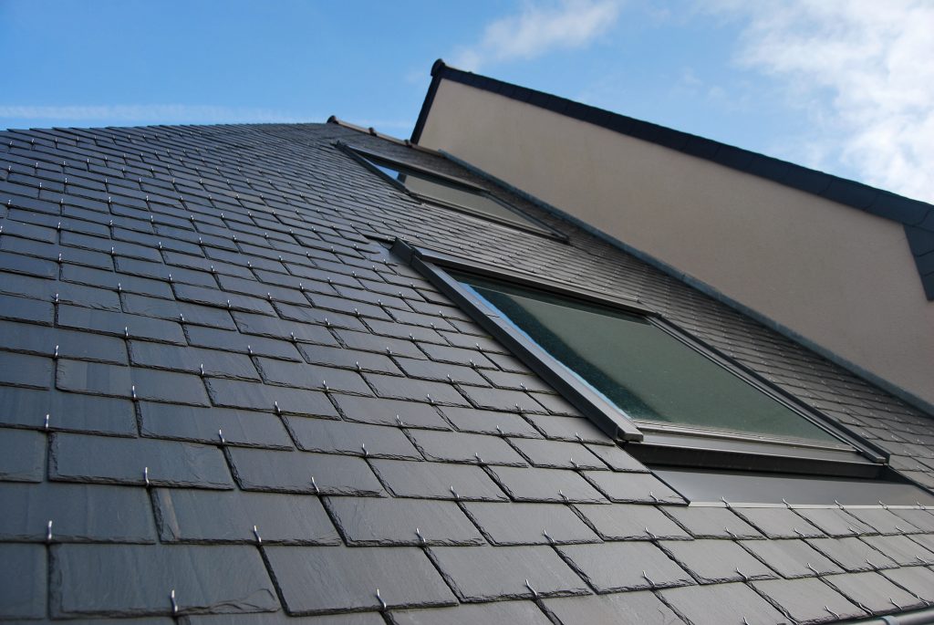 Close-up of a modern building's roof covered with dark slate tiles. Two skylights are visible, set at angles. The sky is clear with a few clouds. The contrasting light and shadow create a dynamic look on the surface.