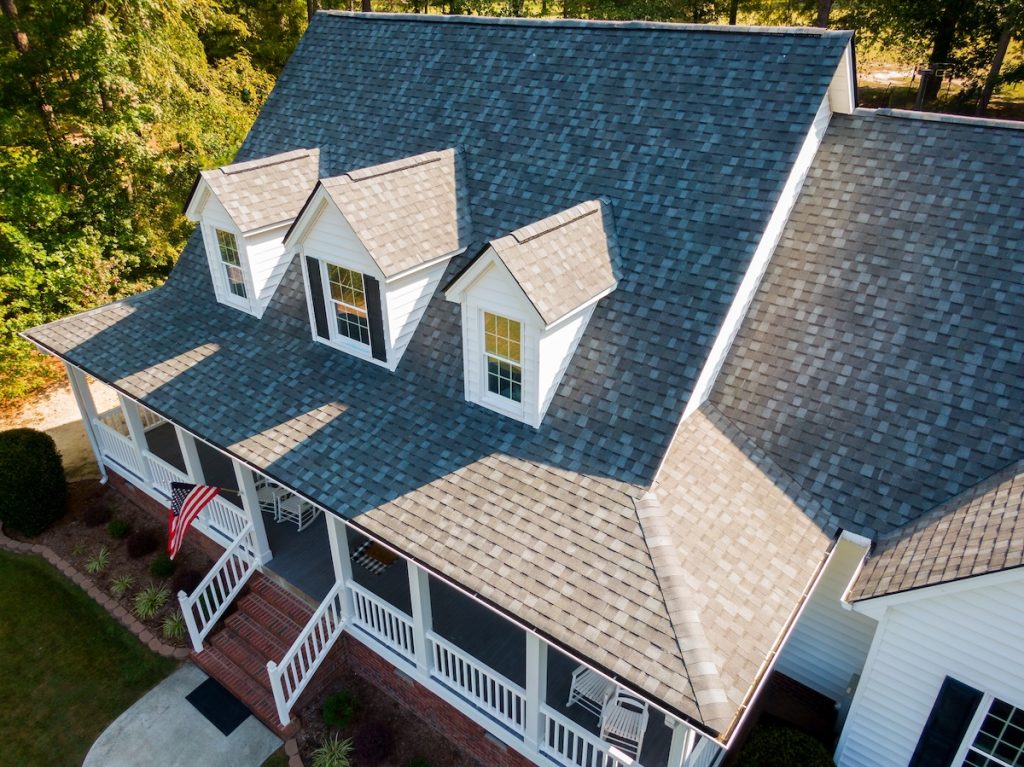 Aerial view of a house with a gray shingle roof, multiple dormer windows, and a large front porch. An American flag hangs by the entrance. The surrounding area has lush greenery.