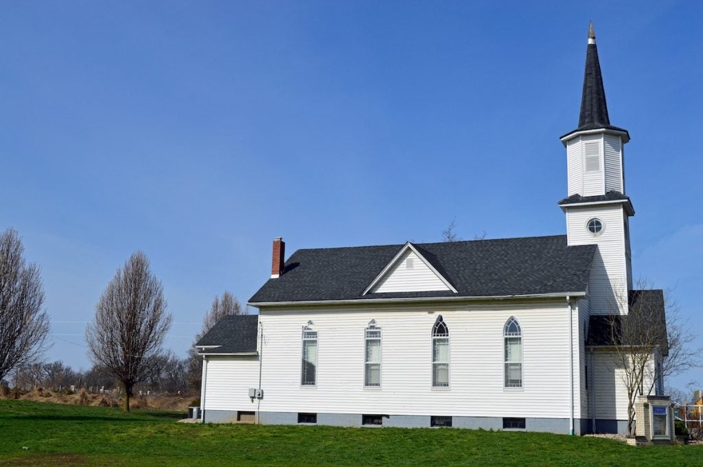 A white church with a tall steeple and arched windows stands on a grassy field. Bare trees surround the building under a clear blue sky.