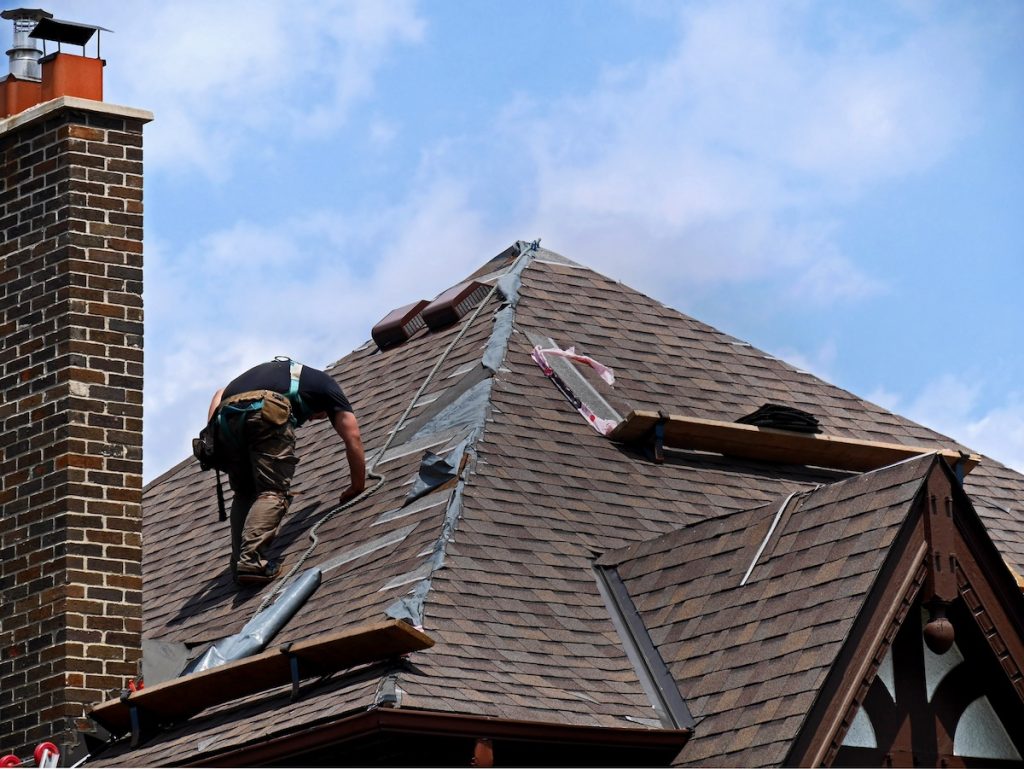 A construction worker repairs a steep, brown shingled roof, surrounded by a chimney and several wooden boards. The sky is partly cloudy, and the worker wears a tool belt and protective gear.
