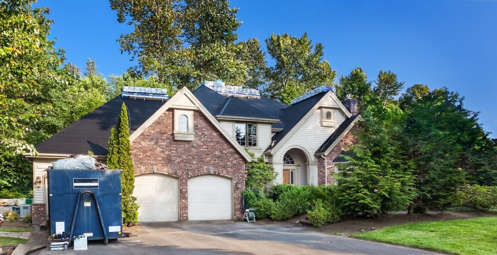 A large, two-story brick house with a steep roof surrounded by trees. Solar panels are visible on the roof, and a trailer is parked in the driveway. The sky is clear and blue, creating a serene atmosphere.