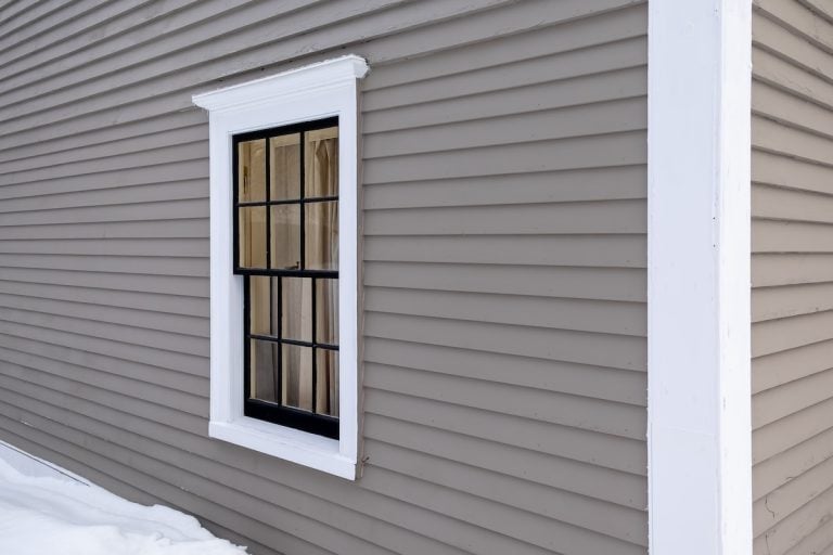 A black-framed window with white trim is set in a gray, horizontally paneled wall. Snow covers the ground below, providing a stark contrast to the building's exterior.