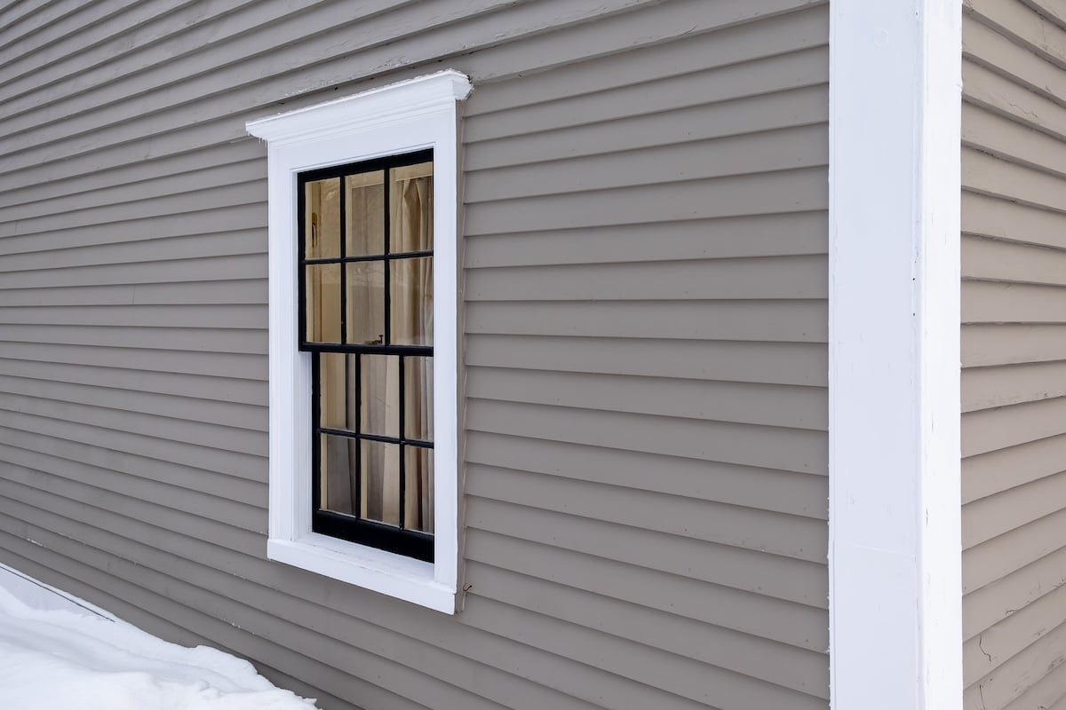 A black-framed window with white trim is set in a gray, horizontally paneled wall. Snow covers the ground below, providing a stark contrast to the building's exterior.