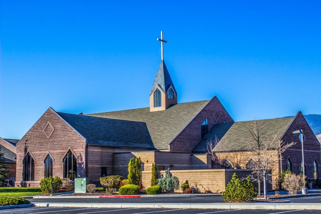 A modern church with a large wooden facade and a steep, pointed roof topped by a cross. It is surrounded by neatly trimmed shrubs and small trees, set against a clear blue sky.