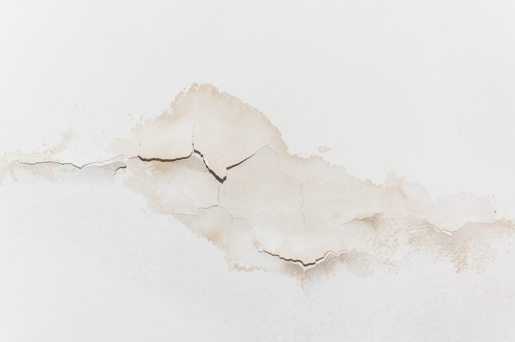 Cracked and peeling paint on a white wall reveals a textured surface beneath, with irregular lines and shades of brown and beige highlighting the damage.