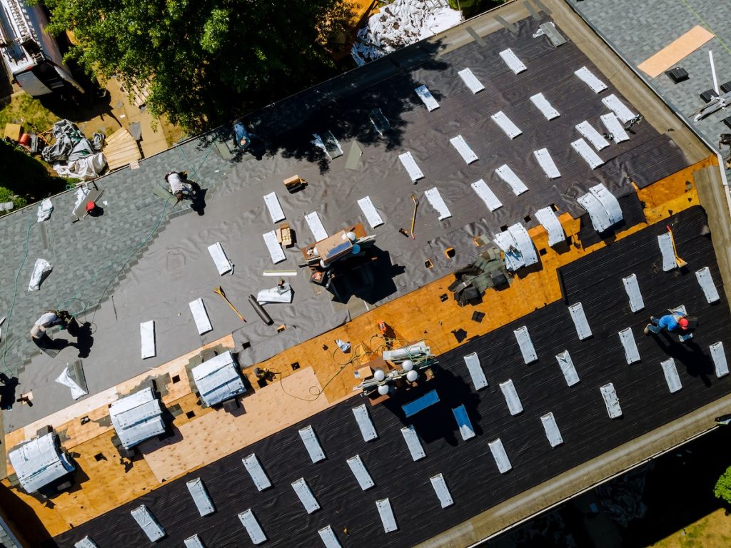 Aerial view of workers installing a new roof on a house. The roof is partially covered with black and gray materials. Various tools and equipment are scattered, and trees are visible nearby.