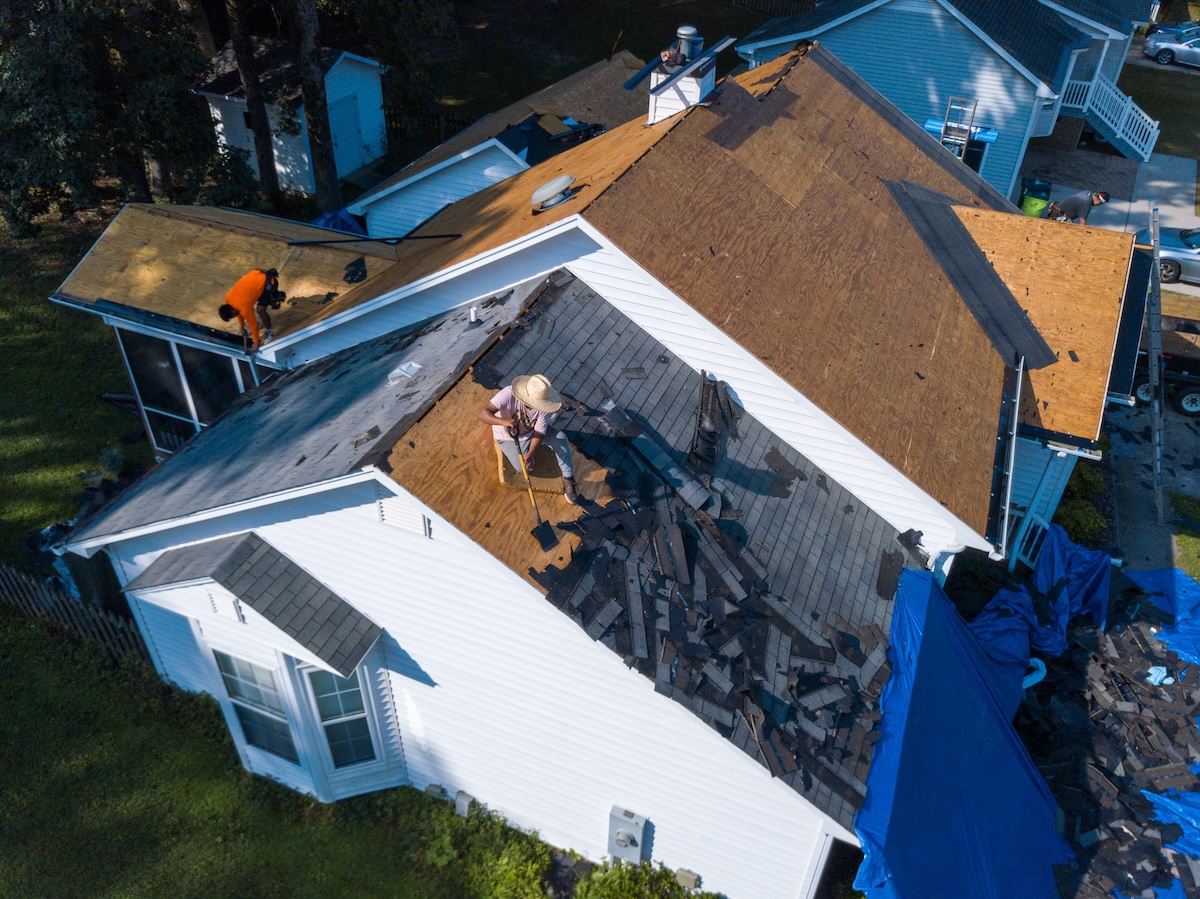 A person wearing a straw hat is working on a roof, removing old shingles. The roof is partially stripped, with debris scattered. Two other people are working on a different section of the roof. The scene takes place in a residential area.