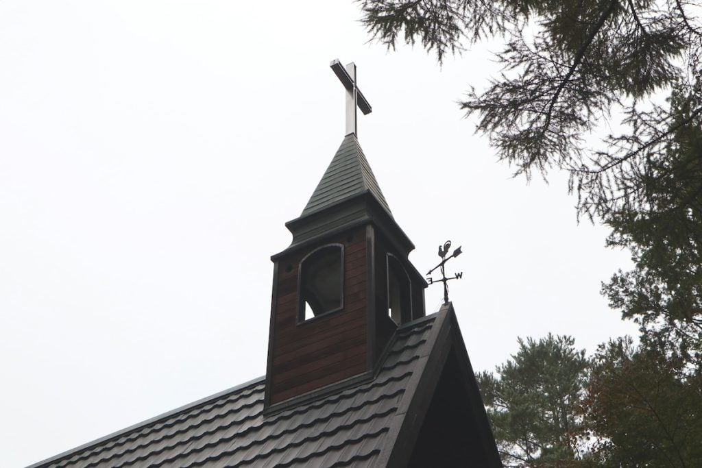 A small chapel with a steep, dark-colored roof topped by a bell tower and cross. A weather vane is visible, pointing towards the sky. Surrounded by trees under a gray, overcast sky.