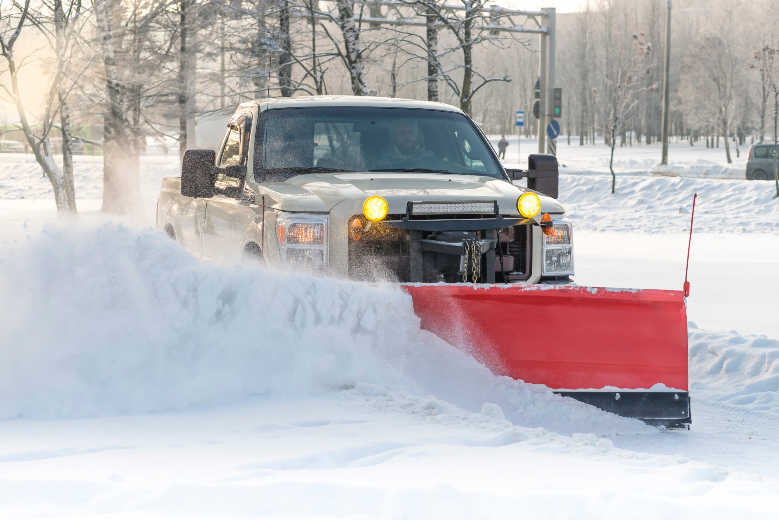 A pickup truck with yellow lights is clearing snow from a road using a large red snowplow on a cold, snowy day. Snow piles up in front of the plow, and trees and traffic signs are visible in the background.