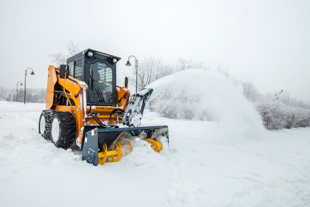 A yellow snow removal vehicle uses a front-mounted snow blower attachment to clear snow in a snowy outdoor area, with trees and streetlights visible in the background.