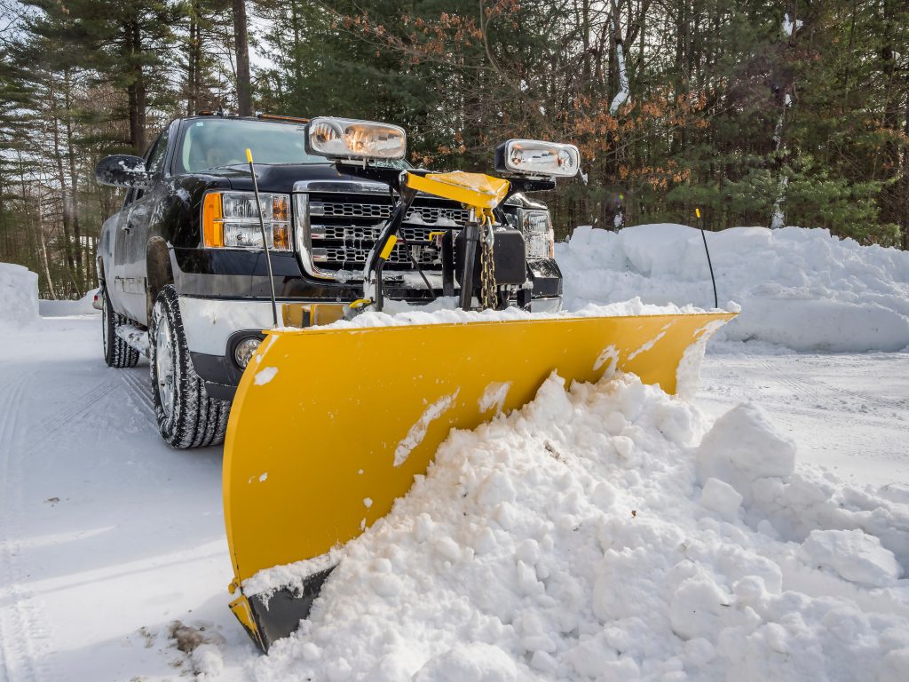 A black pickup truck with a large yellow snow plow attached to the front pushes snow on a snowy road, surrounded by tall piles of snow and trees in the background.