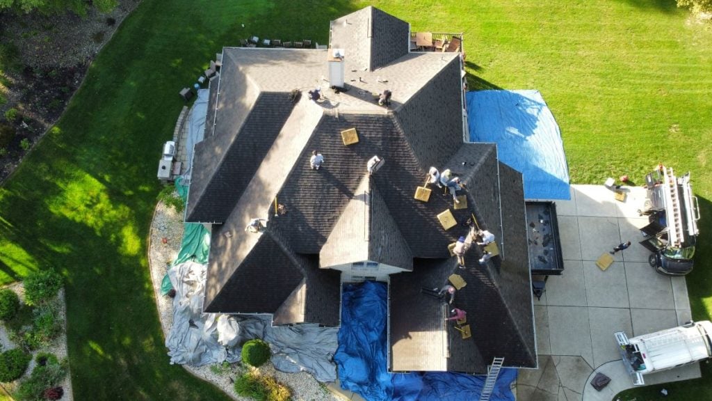 Aerial view of several workers repairing the roof of a large house, with tarps spread on the ground and tools scattered across the roof 