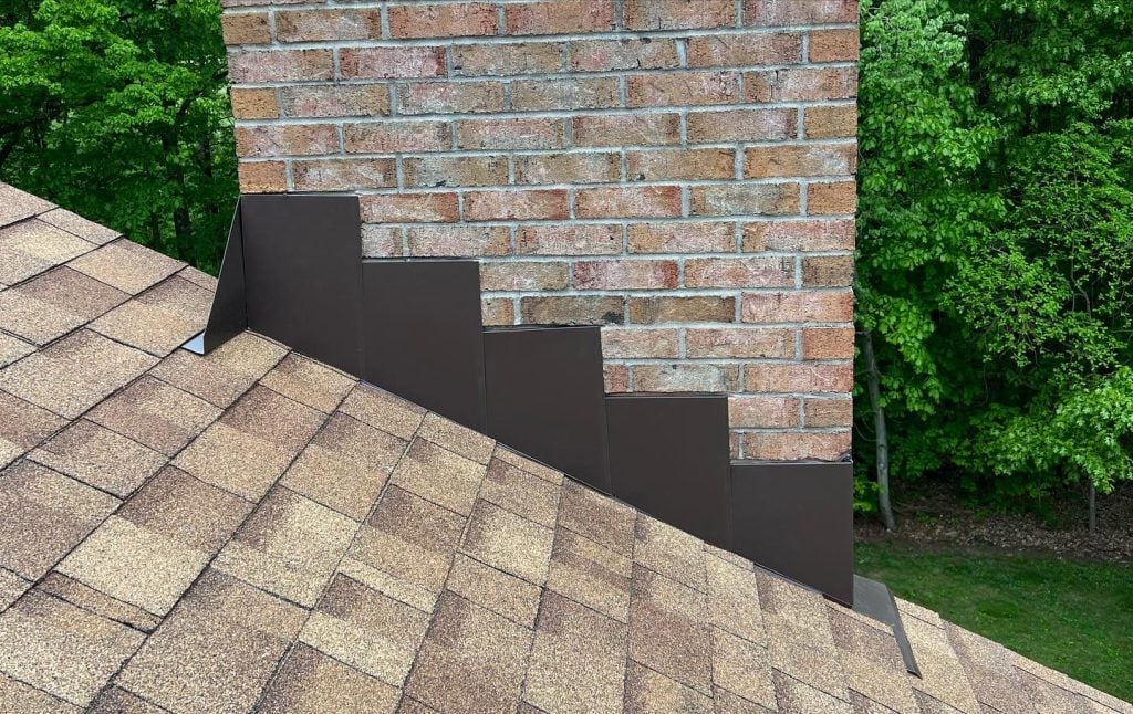 A brick chimney above a brown shingled roof, with dark metal step flashing neatly installed along the side to prevent water leaks