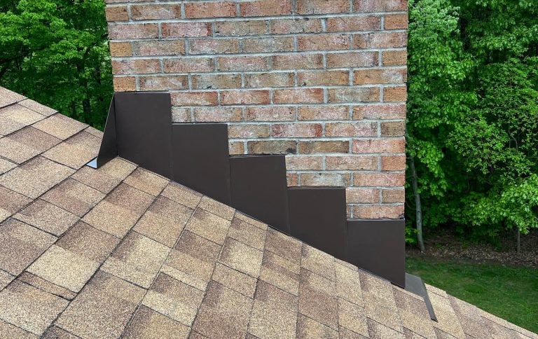 A brick chimney above a brown shingled roof, with dark metal step flashing neatly installed along the side to prevent water leaks