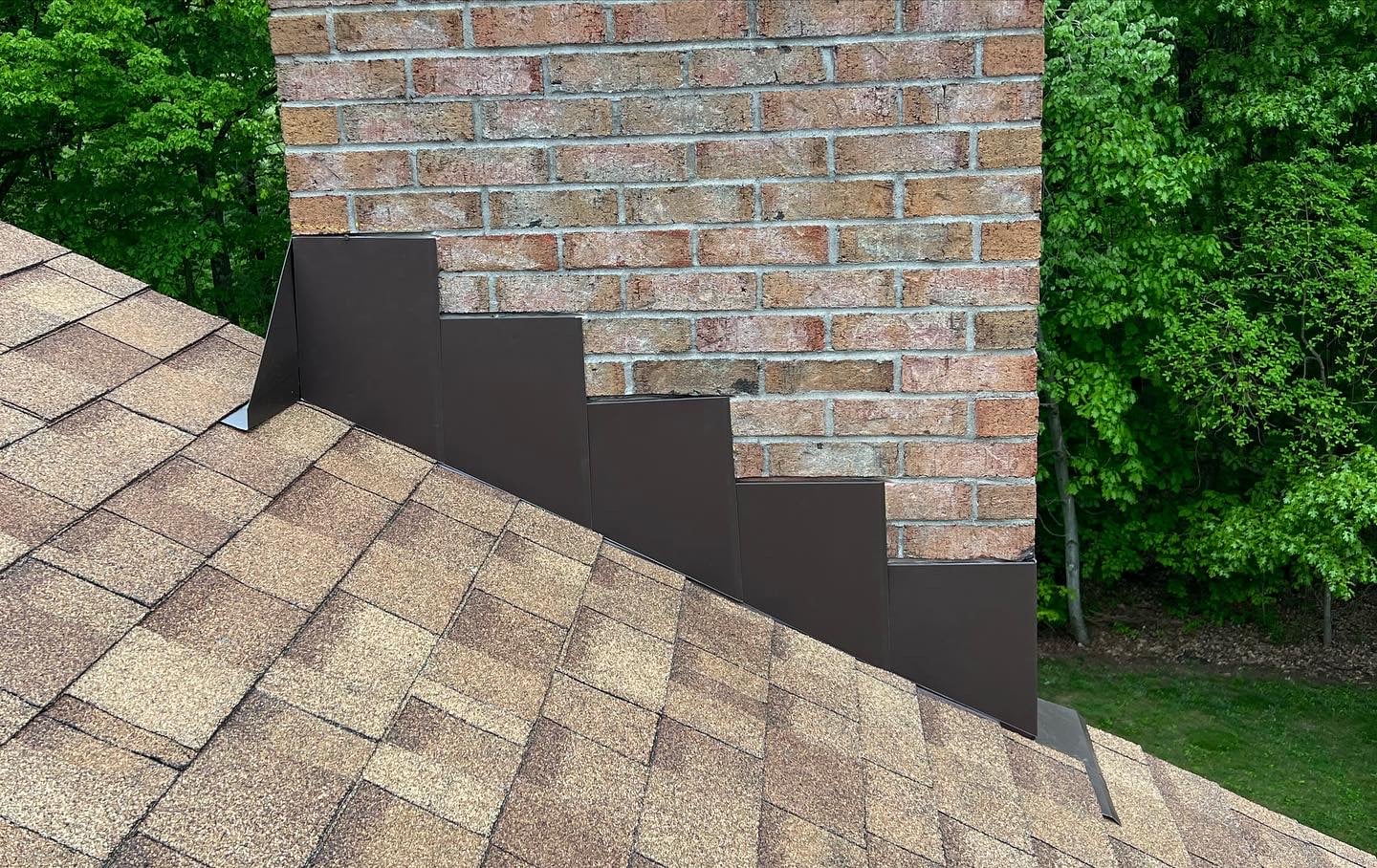 A brick chimney above a brown shingled roof, with dark metal step flashing neatly installed along the side to prevent water leaks