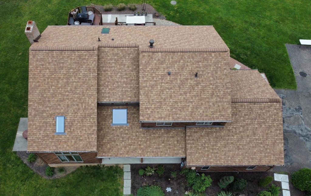 Aerial view of a house with a tan shingle roof by roofing Murrysville, surrounded by green grass, a patio with outdoor furniture, a driveway, and landscaped garden beds.