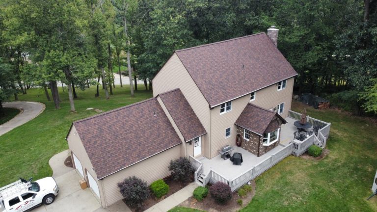Aerial view of a two-story tan house with a brown roof, attached garage, and spacious deck