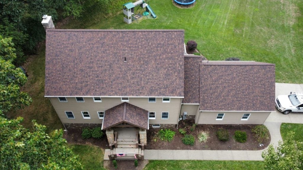 Aerial view of a two-story house with a brown roof, beige siding, and a covered front porch. The yard features green grass, a playset, trampoline, and driveway with a white car—showcasing quality roofing Pittsburgh homes trust.