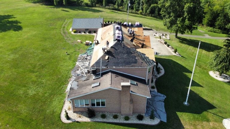 Aerial view of workers replacing the roof of a large brick building surrounded by green grass and trees