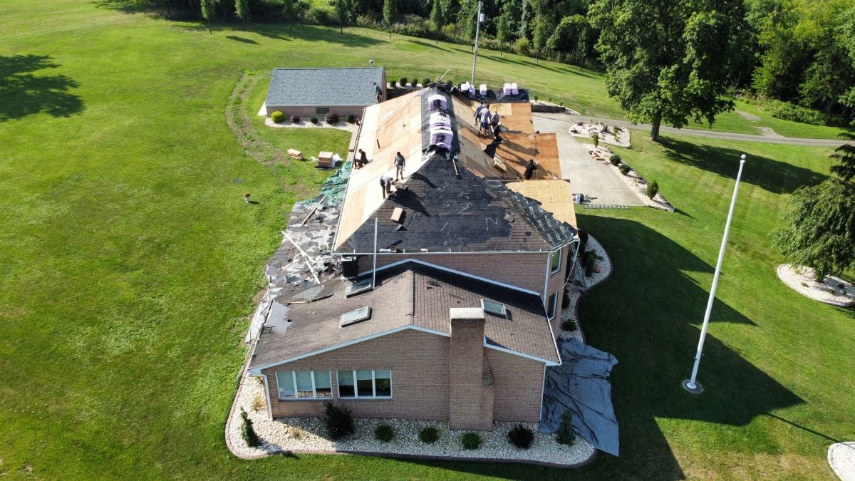 Aerial view of workers replacing the roof of a large brick building surrounded by green grass and trees