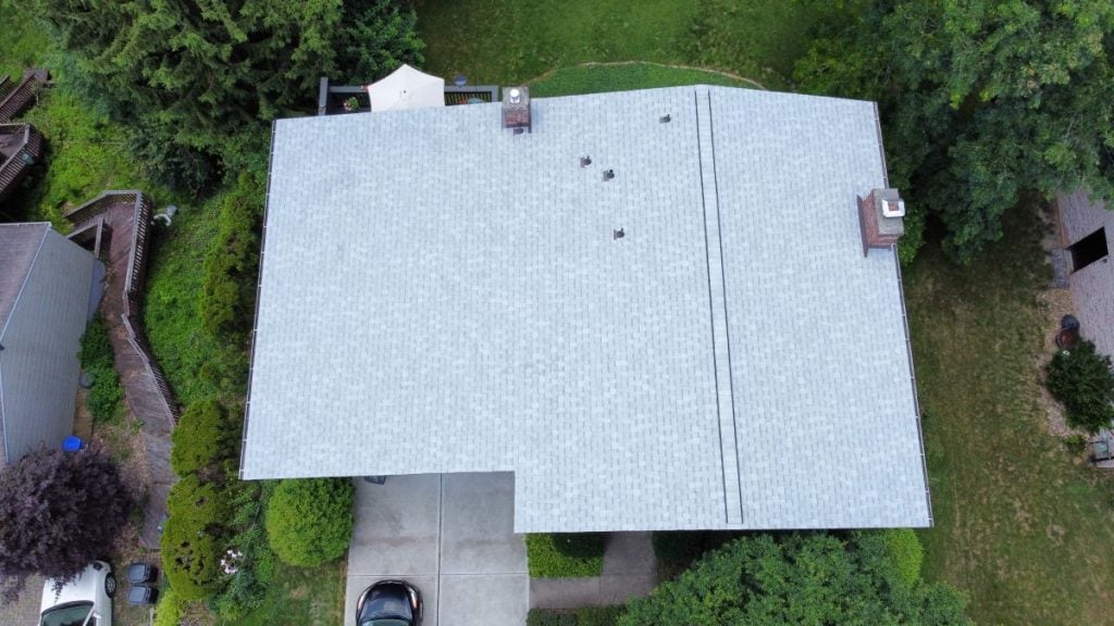 Aerial view of a house with a light gray shingle roof, driveway, and lush greenery.