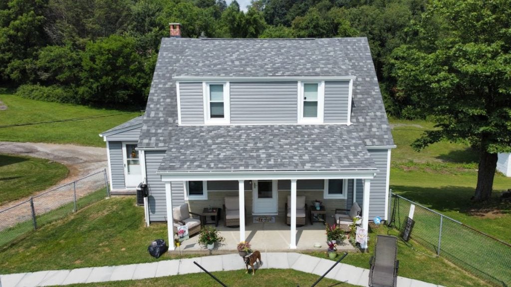 A two-story gray house with a covered front porch, white trim, and a fenced yard showcases quality roofing