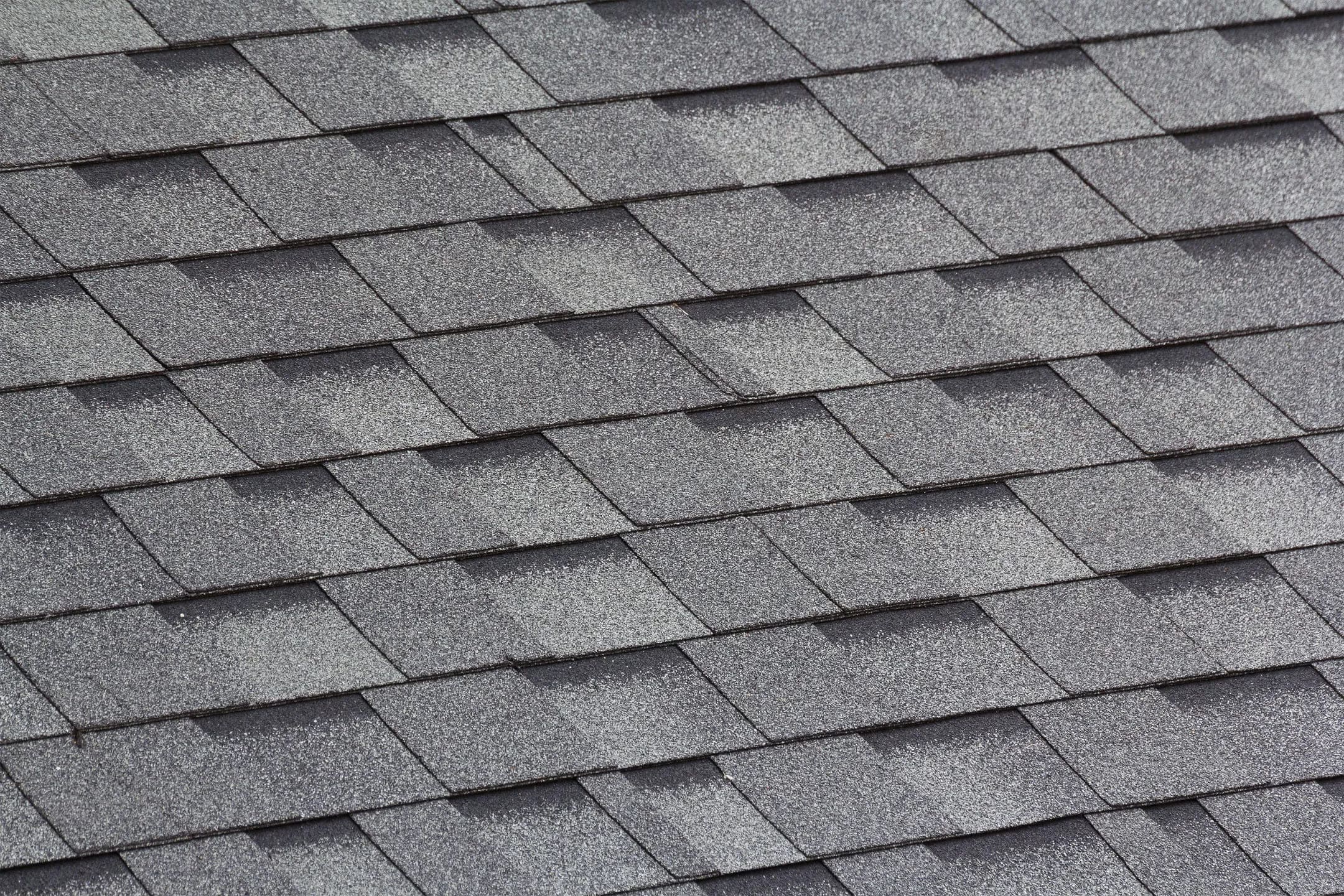 Close-up view of overlapping gray asphalt shingles on a roof, showing the textured surface and regular, repeating pattern of the roofing material.
