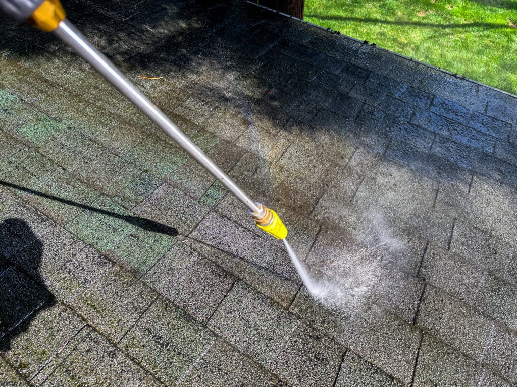 how to clean roof shingles A pressure washer is being used to clean the roof of a house on a clear day. Water sprays over the shingles, effectively removing dirt and debris from the surface